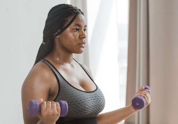 Curvy woman with braided hair working out with dumbbells indoors, embracing body positivity.