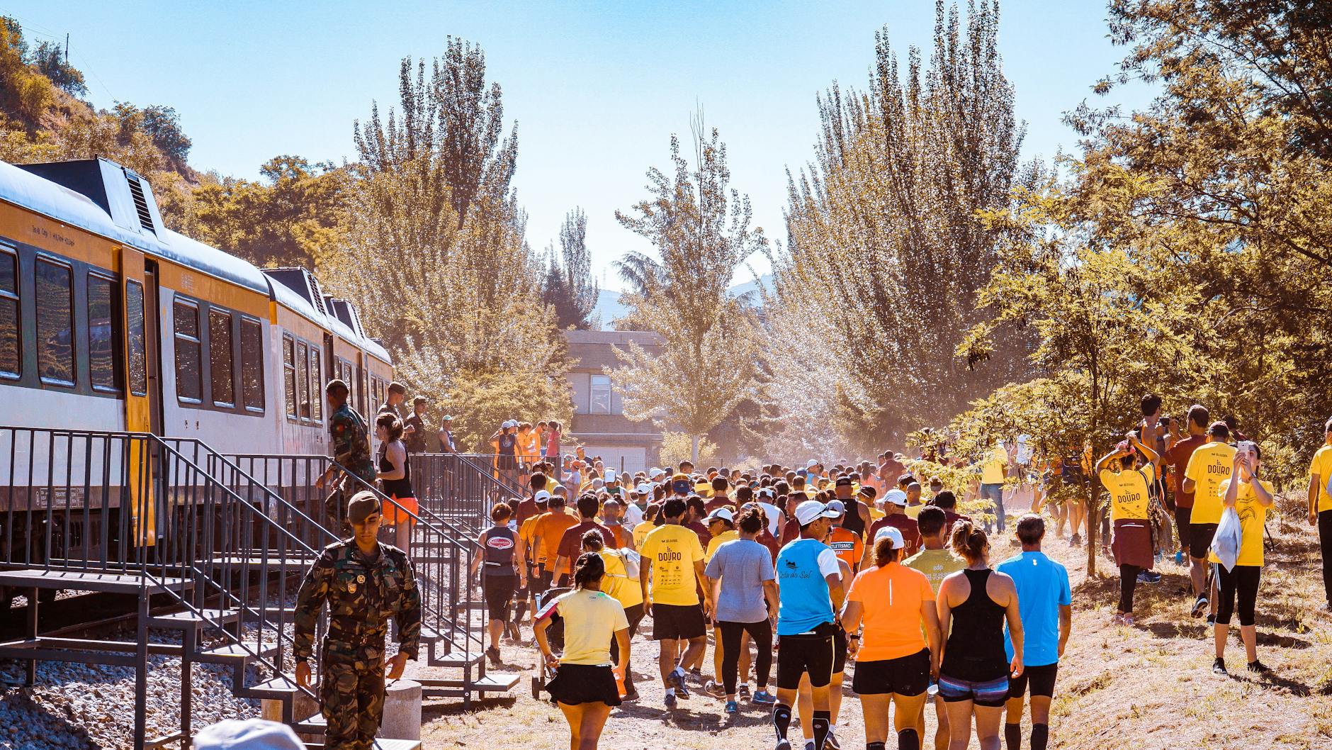 A large group of runners participate in an outdoor event next to a train in a scenic area.