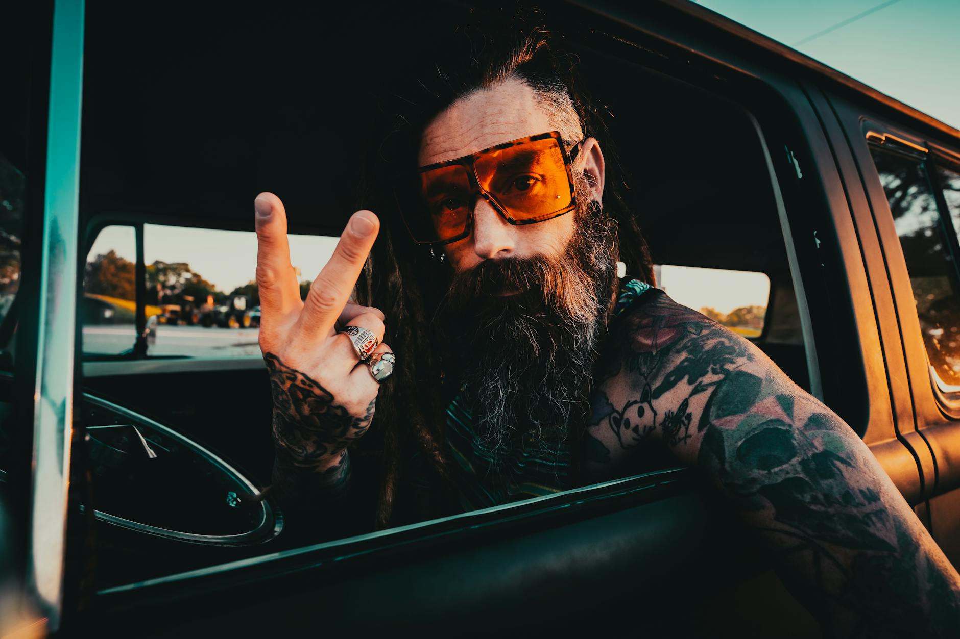 A stylish bearded man with tattoos shows a peace sign while seated in a car with a vintage vibe.