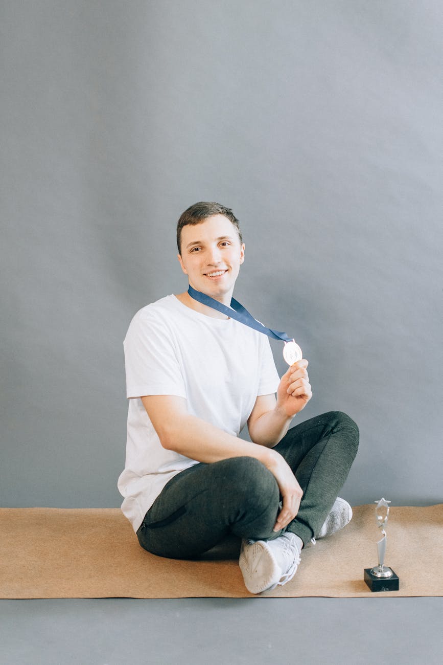 Young man sitting on yoga mat holding medal with a trophy nearby, smiling with pride.