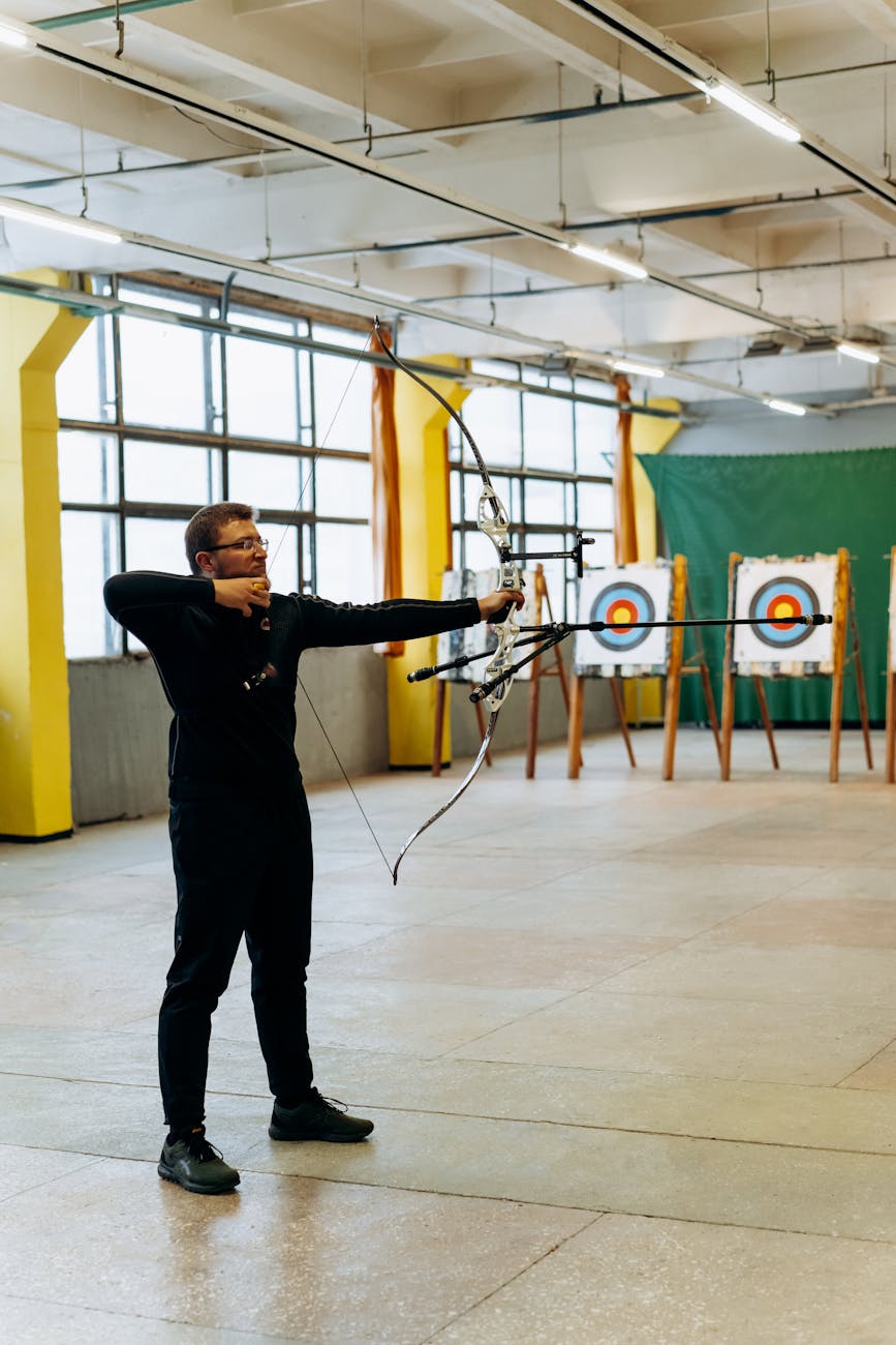 A man practicing archery indoors, aiming at a bullseye target with focused precision.