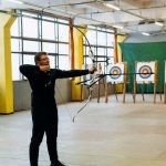 A man practicing archery indoors, aiming at a bullseye target with focused precision.