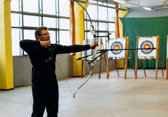 A man practicing archery indoors, aiming at a bullseye target with focused precision.
