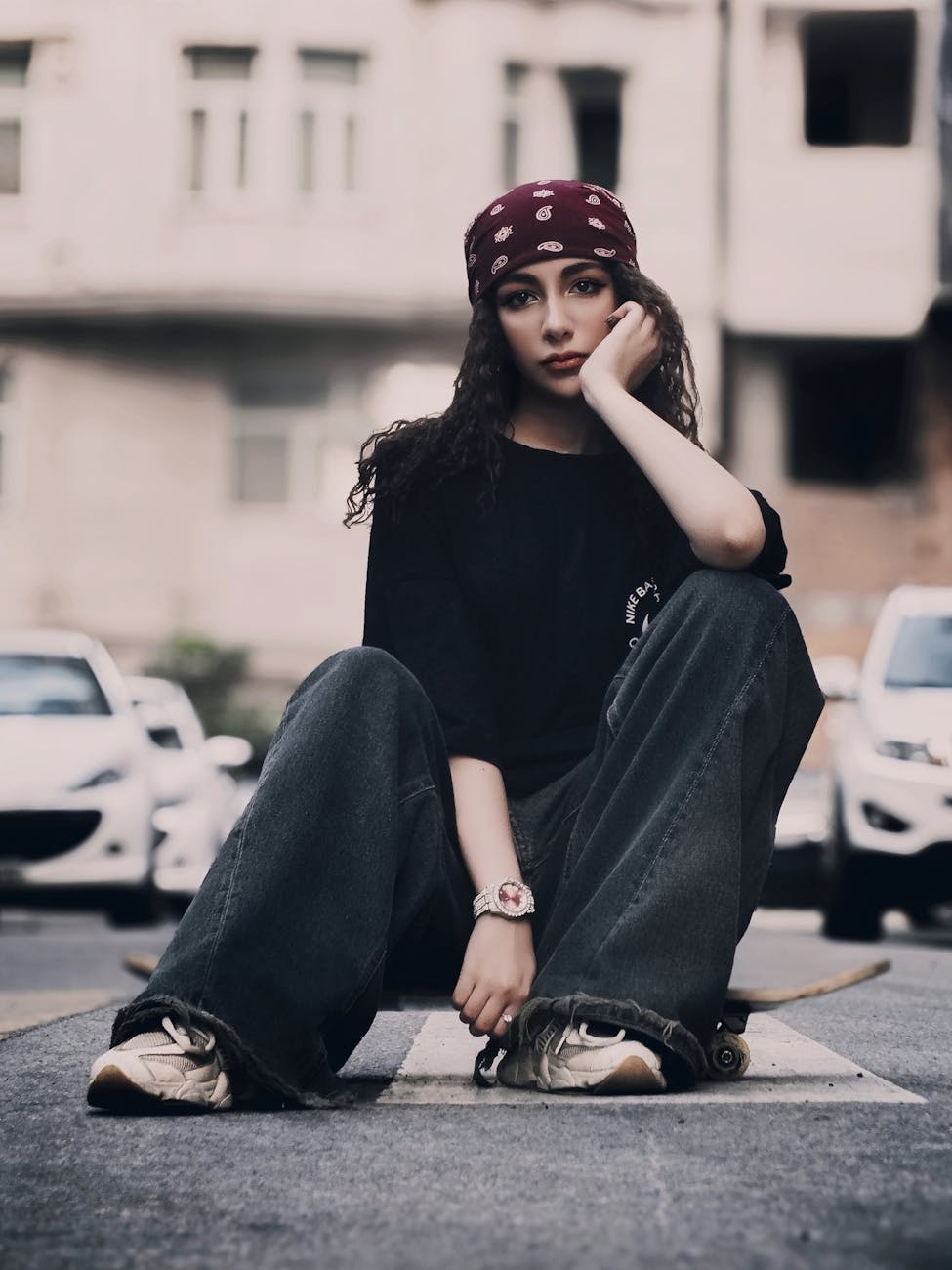 Stylish young woman in casual attire sitting on a skateboard on a city street.