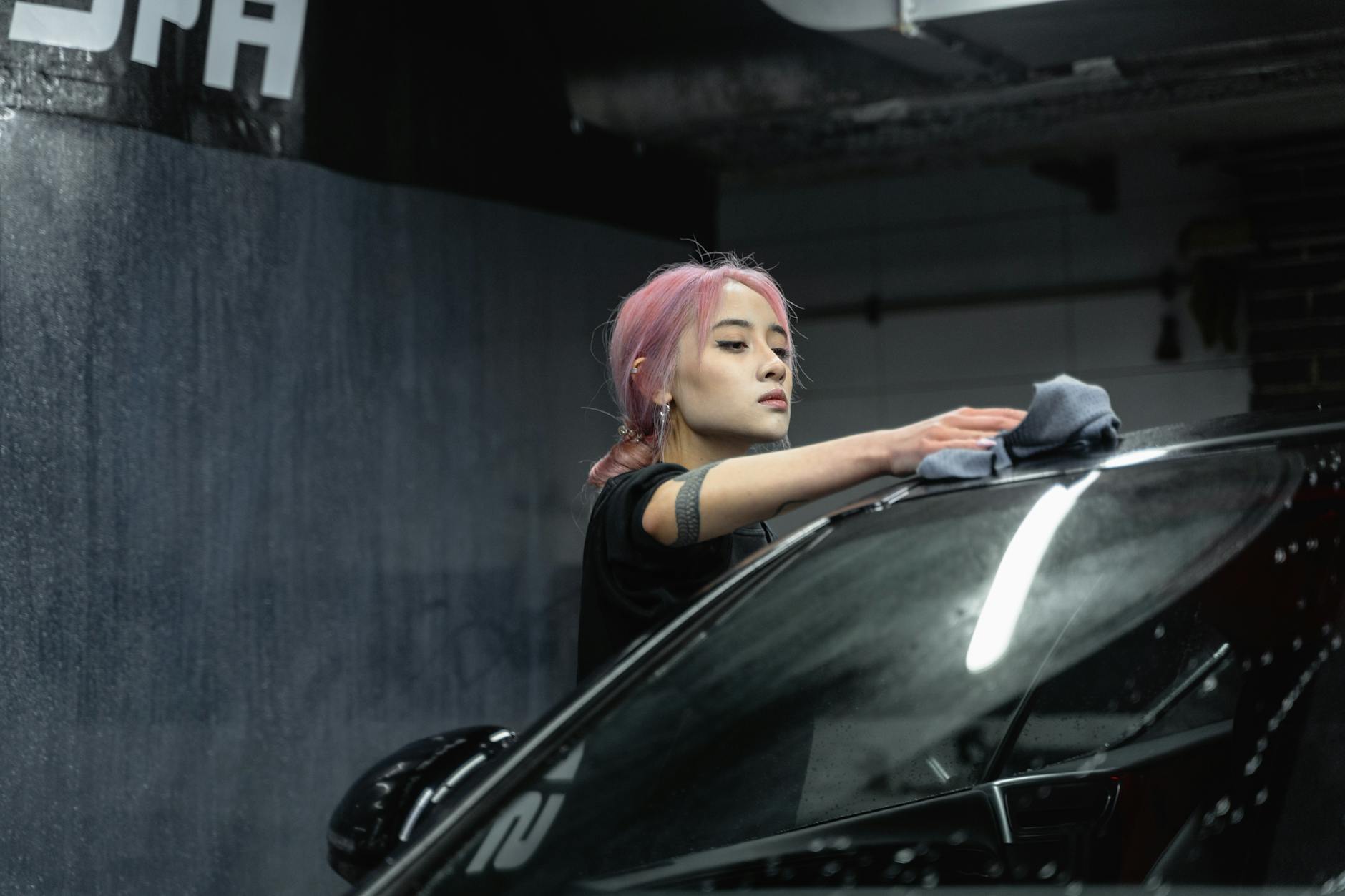 A young woman with pink hair wipes a car windshield with a microfiber cloth indoors.