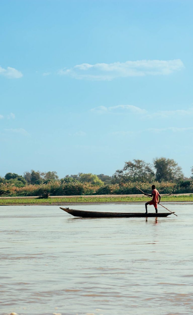 A serene scene of a man paddling a traditional wooden boat on a calm river under a clear blue sky.