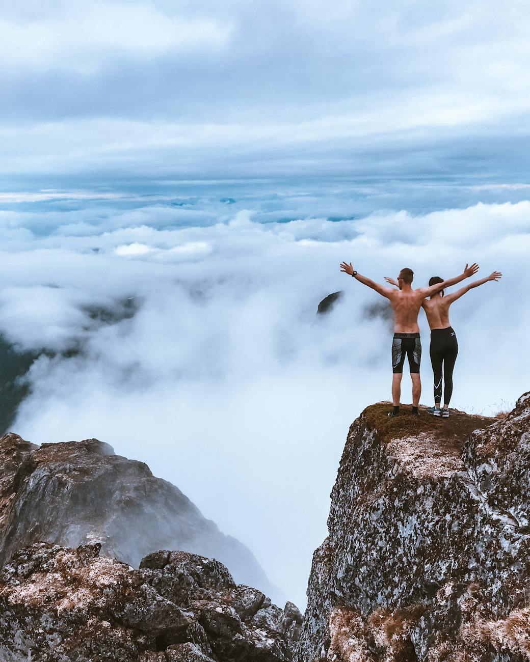 Two hikers stand on a rocky cliff, embracing the expansive mountain view and clouds, symbolizing freedom and exploration.