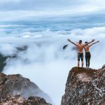 Two hikers stand on a rocky cliff, embracing the expansive mountain view and clouds, symbolizing freedom and exploration.