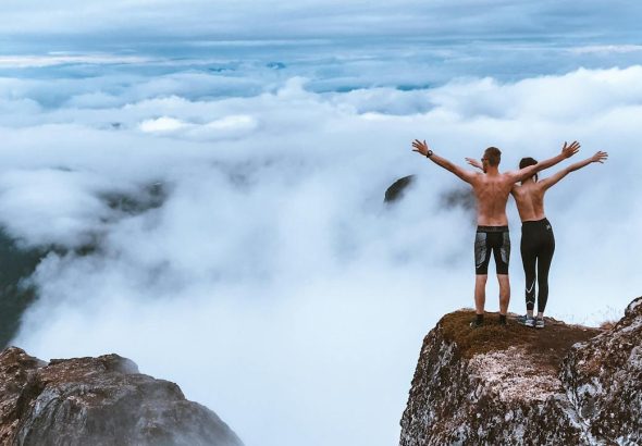 Two hikers stand on a rocky cliff, embracing the expansive mountain view and clouds, symbolizing freedom and exploration.